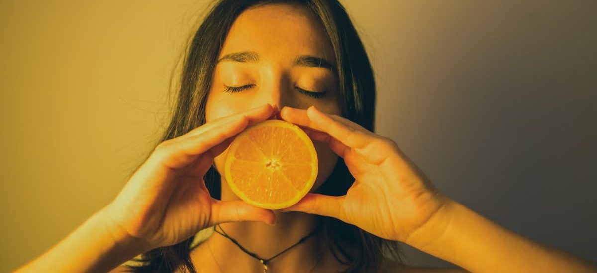 Calm young female with bare shoulders standing with eyes closed against plain wall and covering lips with orange slice