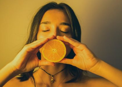 Calm young female with bare shoulders standing with eyes closed against plain wall and covering lips with orange slice