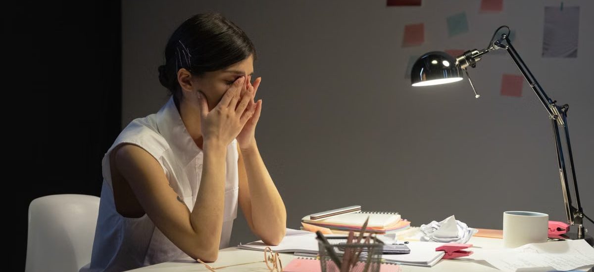 A stressed businesswoman rubs her eyes at a cluttered desk, overwhelmed by work.