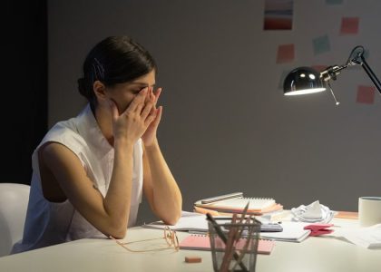 A stressed businesswoman rubs her eyes at a cluttered desk, overwhelmed by work.